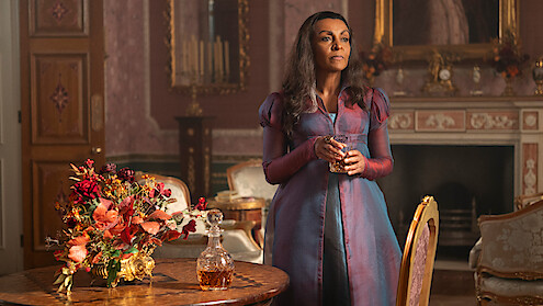 Woman in a purple Regency dress stands by a table with flowers and a decanter in an ornate, classic room with a fireplace, mirror, and rich decor, holding a glass and looking thoughtful.