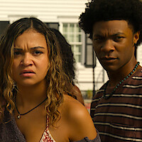 Two young adults stand outdoors in daylight, looking concerned. They are in front of a white house with windows and shutters, suggesting a residential neighborhood setting.