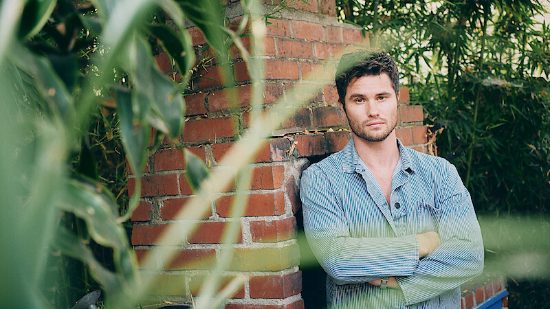 A man in a light denim shirt stands with arms crossed in front of a brick wall outdoors, surrounded by green plants and bamboo. The setting is natural and relaxed, with leaves partially obscuring the foreground.