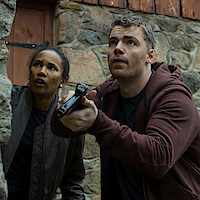 Two people outside by a stone wall, looking alert and cautious. One holds a handgun, both in casual clothing, suggesting a tense or dangerous situation, possibly investigating or preparing for action.