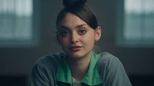 Young woman with dark hair in a bun, wearing a green-collared gray shirt, sitting indoors in a softly lit room with large windows in the background, looking directly at the camera with a calm expression.
