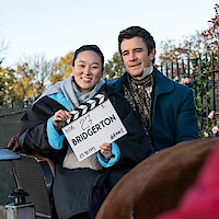 Two people sitting outdoors on a film set dressed in period costumes, one holding a Bridgerton clapperboard, with a fence, flowers, and trees in the background.
