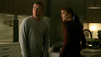 A man and a woman stand in a modern, dimly lit kitchen, both looking slightly off-camera with serious expressions, surrounded by contemporary decor and appliances.