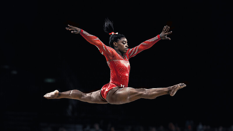 Gymnast Simone Biles doing a split leap in the air, wearing a red leotard.