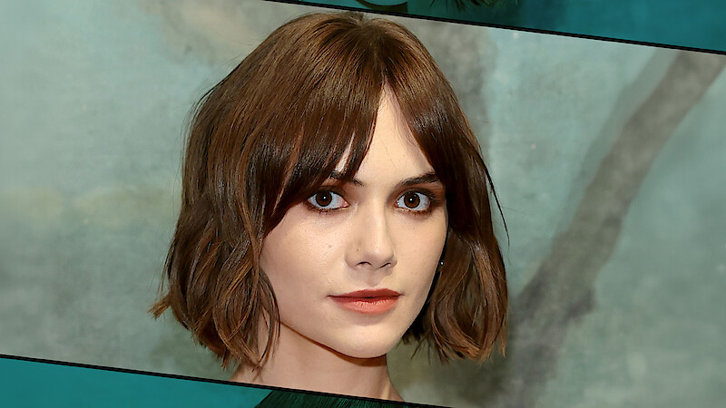 Woman with short brown hair and bangs stands in front of a soft, painted blue-green background with tree details, looking directly at the camera, in a close-up portrait setting.
