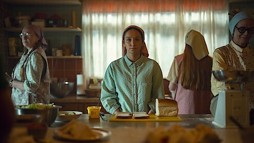Young woman stands at a kitchen counter with a loaf of bread, surrounded by three women preparing food in a warmly lit, vintage-style kitchen with curtains and shelves in the background.
