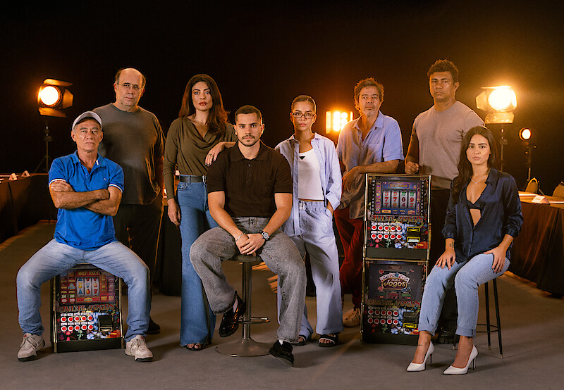 Elenco principal da série "Donos do Jogo" posa ao lado de máquinas caça-níquel