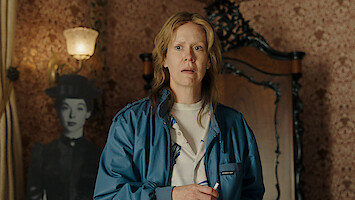 A concerned person in a blue jacket stands in a vintage room with patterned wallpaper, ornate wooden furniture, and a black-and-white cutout of a woman in period clothing in the background.