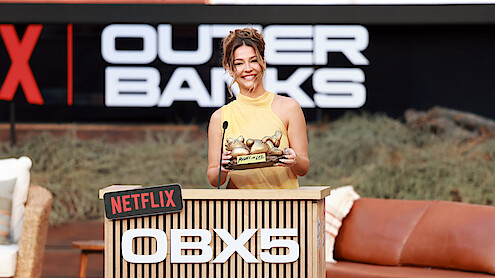 Smiling woman in yellow dress holding award, standing at a Netflix OBX5 podium outdoors with Outer Banks signage and casual seating in the background.