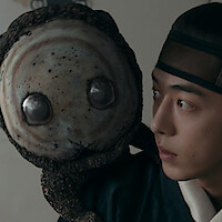 A young man in traditional Asian attire looks at a round, textured creature with large glassy eyes perched on his shoulder, indoors with soft lighting.