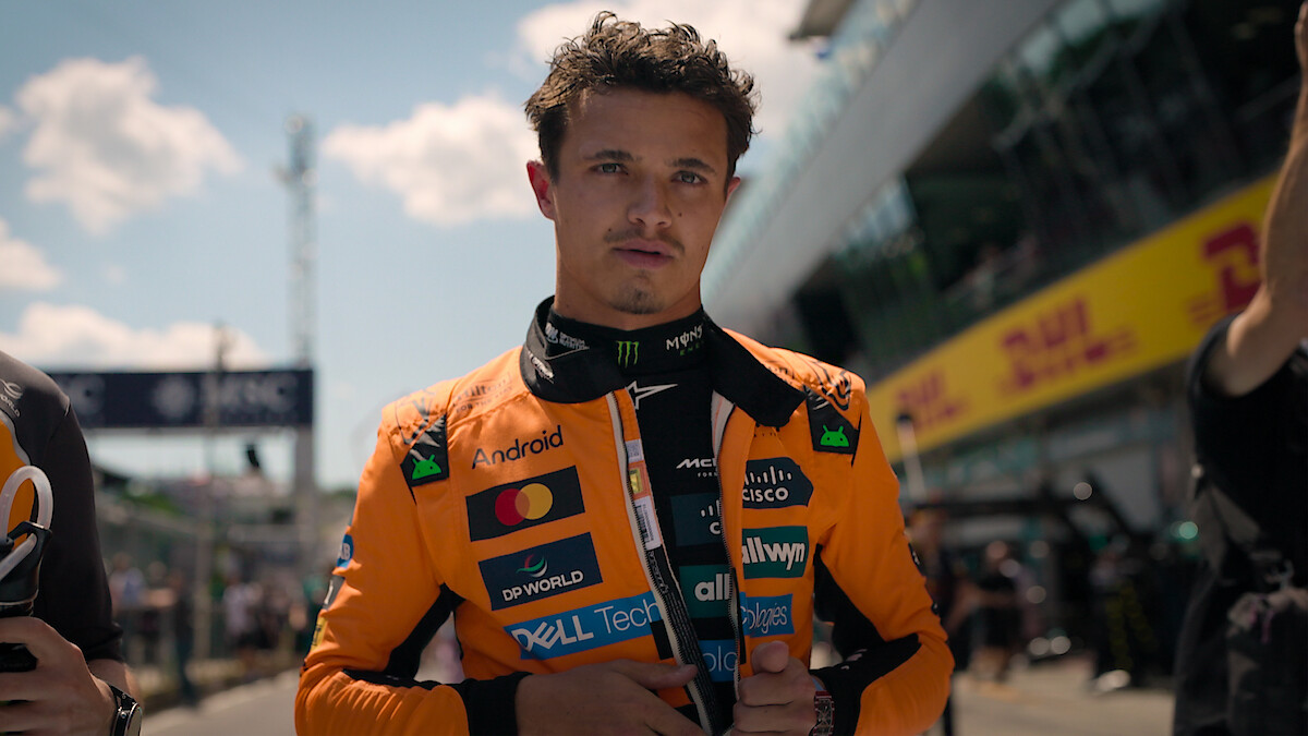 Lando Norris in an orange racing suit stands on a sunlit pit lane, surrounded by team members and blurred race track environment, preparing for a race.