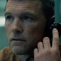 Close-up of a man with short hair and stubble holding a black telephone receiver to his ear, appearing thoughtful. He is indoors, possibly in a serious or intense setting with muted lighting and a blue background.