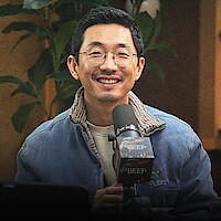 Lee Sung Jin sits indoors in front of a microphone and laptop, with green plants in the background, appearing to host a podcast.