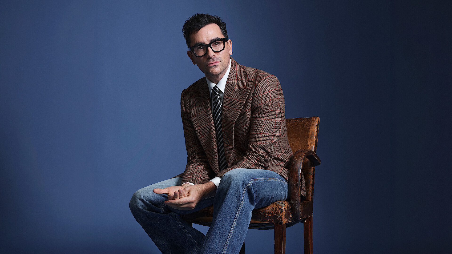Dan Levy sits in a chair as he poses against a blue backdrop, wearing a checked blazer.