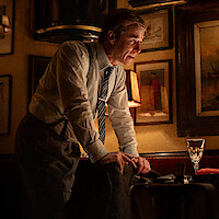 A man in formal attire leans over a restaurant table in a dimly lit, vintage-style room decorated with framed paintings and warm lighting, creating a classic and intimate atmosphere.