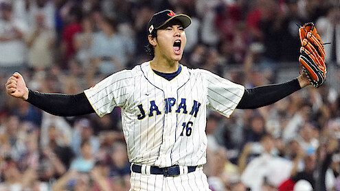 Shohei Ohtani celebrating on the field, arms raised and mouth open in excitement, wearing a white pinstripe jersey and glove, with a blurred crowd in the stadium background.