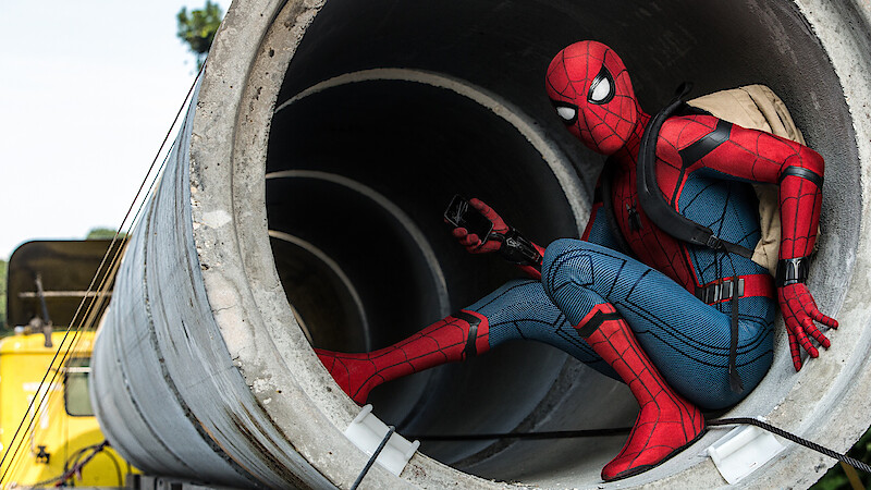 A person in a Spider-Man costume crouches inside a large concrete pipe outdoors, holding a device and wearing a backpack, with construction vehicles and trees visible in the background.