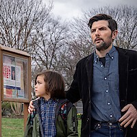 A concerned man with a beard stands outdoors with a young child, both looking in the same direction. Behind them is a wooden bulletin board with "Have you seen me?" posters. Leafless trees and cloudy sky suggest winter or early spring.