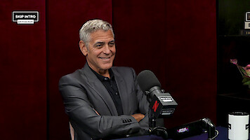 A man in a dark suit sits smiling at a podcast studio table, with a large microphone and "Skip Intro" branding, set against a red background. The mood is relaxed and professional.