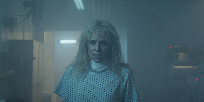 Karen Wheeler, her blonde hair disheveled, stands in a dim, hazy hospital room, wearing a medical gown amid harsh lighting and looming industrial equipment.