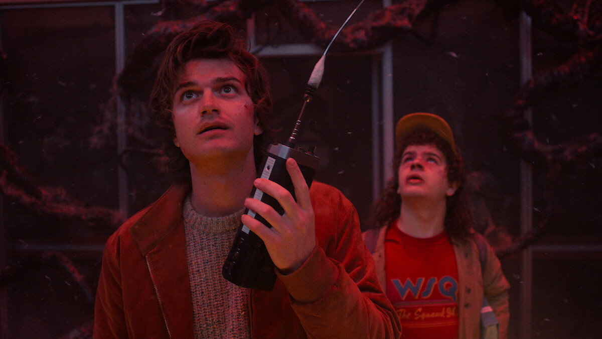 Steve and Dustin in an eerie environment with red lighting, holding a walkie-talkie and looking upward with concerned expressions as mysterious vines cover the background in 'Stranger Things 5'