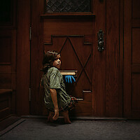 A young child kneels inside a dimly lit house by a wooden door, turning to look back as a blue-gloved hand reaches through the mail slot, creating a mysterious and suspenseful atmosphere.