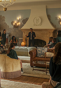 A group of people gathers in a luxurious living room with ornate furniture and a large decorative fireplace, as a man stands and speaks to the seated guests in a warmly lit, elegant environment.