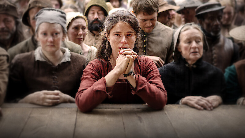 A distressed young woman in historical clothing sits at a wooden table, hands clasped, surrounded by somber villagers in period attire, suggesting a dramatic or tense moment in a historical community setting.