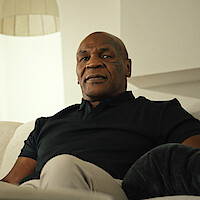 Man sitting on a light-colored couch in a modern, softly lit living room, wearing a dark shirt and light pants, looking relaxed.