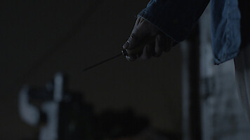 A person holding a knife in their right hand at night, wearing a dark blue jacket. The background is dimly lit, creating a suspenseful or tense atmosphere.