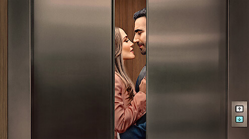 A man and woman standing closely, facing each other romantically inside an elevator as the doors begin to close, suggesting a private or intimate moment in a modern building setting.