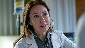 A female doctor wearing a white coat and stethoscope listens attentively to a patient in a hospital room with medical equipment visible in the background.