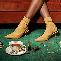 Woman’s legs in yellow heels and socks on green carpet, vintage teacup with lipstick mark, gold necklace, lipstick, cozy retro setting with red sofa background.
