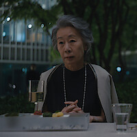 Youn Yuh-jung as Chairwoman Park sits at an outdoor dining table at night, in front of a glass of white wine and a modern meal, with trees and city lights in the background.