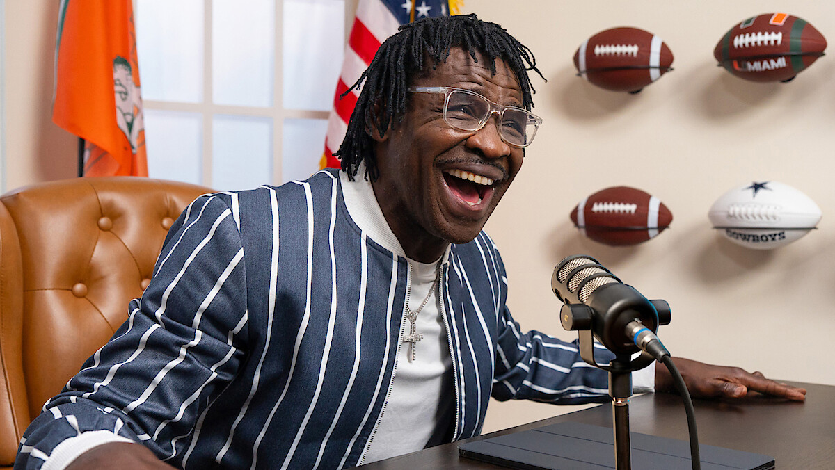 Man in striped jacket smiling and speaking into a microphone at a desk, with footballs mounted on the wall and flags in the background, resembling a sports interview or podcast setting.