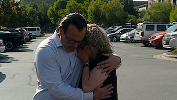 Two people embrace emotionally in a parking lot surrounded by cars and trees, suggesting comfort or consolation in a public, outdoor setting on a sunny day.