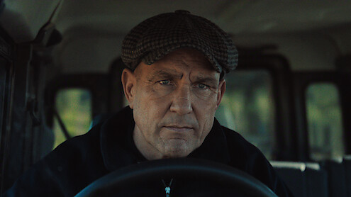 Middle-aged man wearing a flat cap sits in the driver’s seat of a vehicle, hands on the wheel, looking seriously forward. The interior is dimly lit, and trees are visible through the windows in the background.