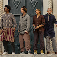 Four young adults in casual, loose clothing stand on stone steps in front of large ornate doors, looking alert and concerned, with one holding a handgun; the environment appears historic or ancient.