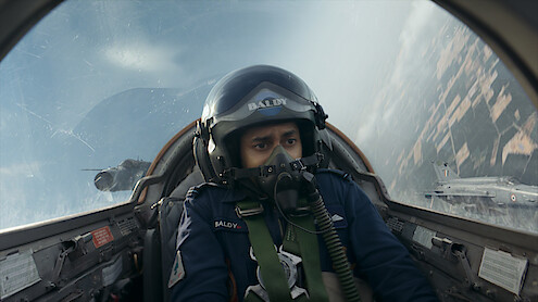 Pilot in cockpit flying a jet fighter, wearing helmet and oxygen mask, with intense expression; another jet visible outside, sky and landscape blurred from high speed and aerial maneuver.