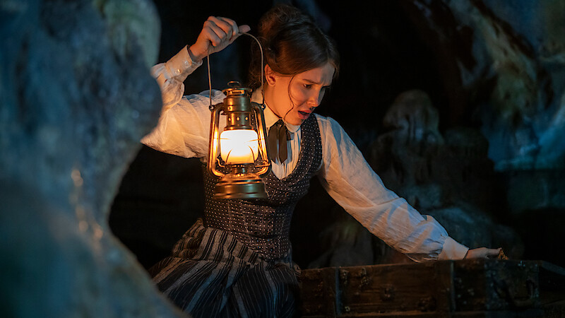 A woman in period clothing holds a lit lantern while exploring a dark, rocky cave, leaning over an old wooden chest, suggesting a search or discovery in a mysterious setting.