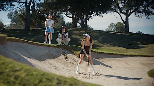 Mikaela Hoover as Ava prepares to hit a golf ball from a sand bunker as two people watch nearby on a sunny golf course surrounded by trees and grass.