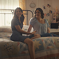 Two women sitting on a bed in a cozy, sunlit bedroom decorated with photos, a laptop, and floral curtains, smiling and enjoying a relaxed moment together.
