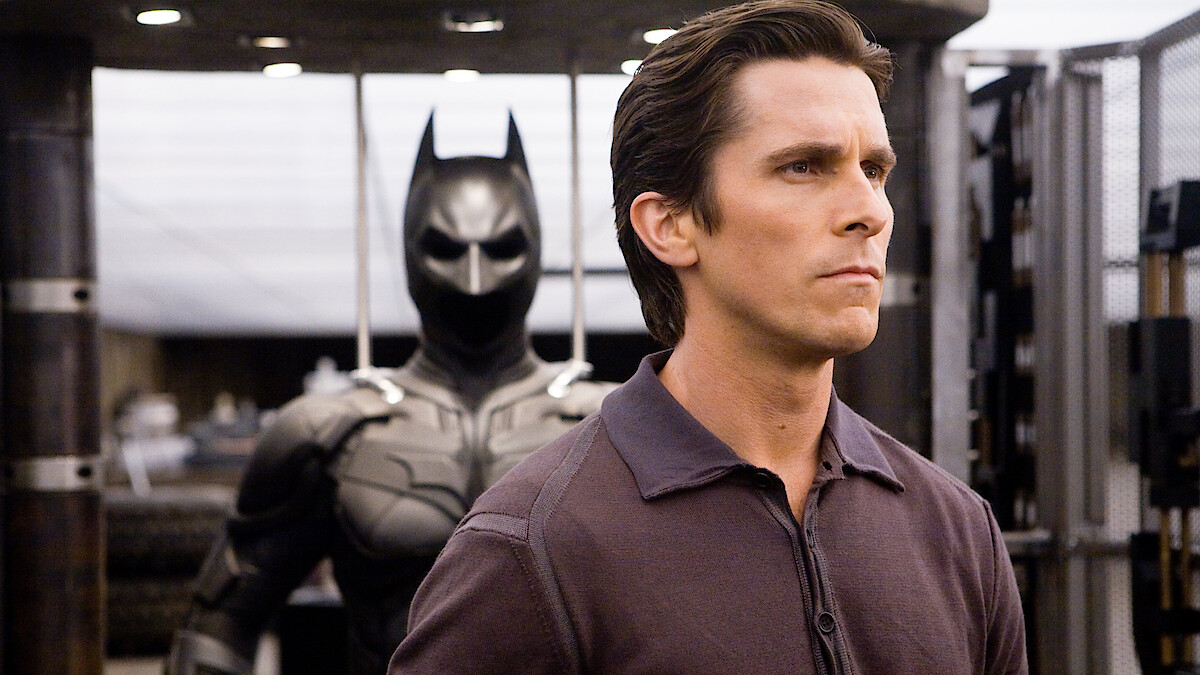 Christian Bale stands in front of Batman costume in The Dark Knight.