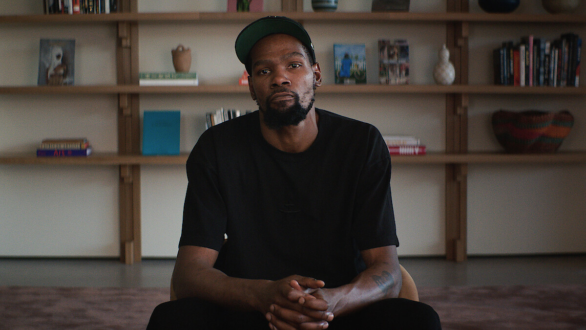 A man in a black shirt and cap sits indoors with hands clasped, in front of a bookshelf filled with books, vases, and decorative objects, giving a calm, introspective mood.