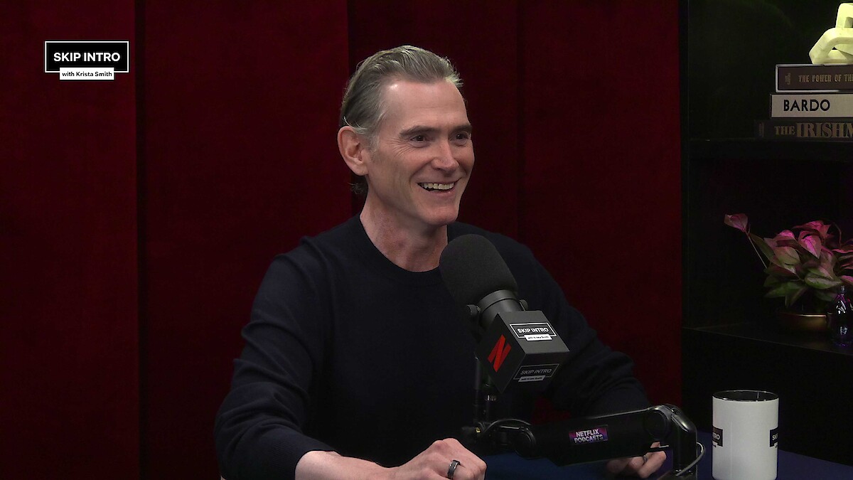 A smiling man sits in a podcast studio with a microphone, dark red curtains, and shelves with books and flowers behind him. The mood is relaxed and conversational, suggesting a friendly interview setting.