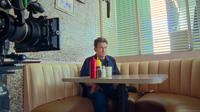 Man sitting alone in a retro diner booth with condiments on the table, film camera and boom microphone visible, sunlight filtering through blinds.