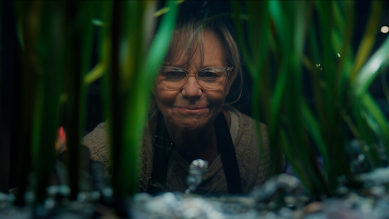 Sally Field as Tova wearing glasses looking into an aquarium tank, viewed through tall green aquatic plants and aquarium decorations, with a focused expression in a dim indoor setting.