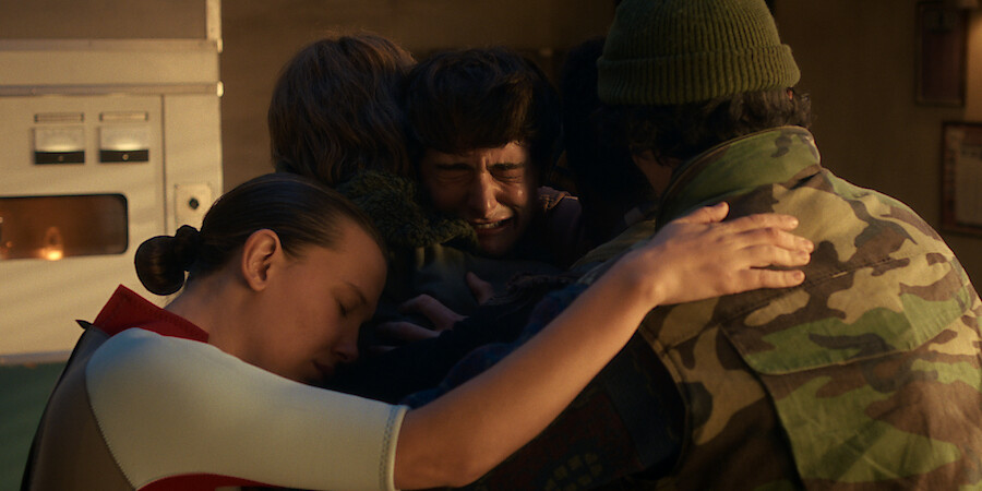 Eleven, Jonathan Byers, Will Byers, and Mike Wheeler sharing an emotional group hug indoors, with eyes closed, in a warmly lit room.
