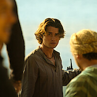 A young man with wavy hair stands outdoors near water, holding a walkie-talkie, looking toward the camera, surrounded by people dressed in casual clothing under warm sunlight.