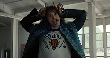 Eddie Munson (Joseph Quinn) stands indoors, playfully sticking out his tongue and making horn gestures with his hands while wearing a "Hellfire Club" t-shirt and denim jacket.